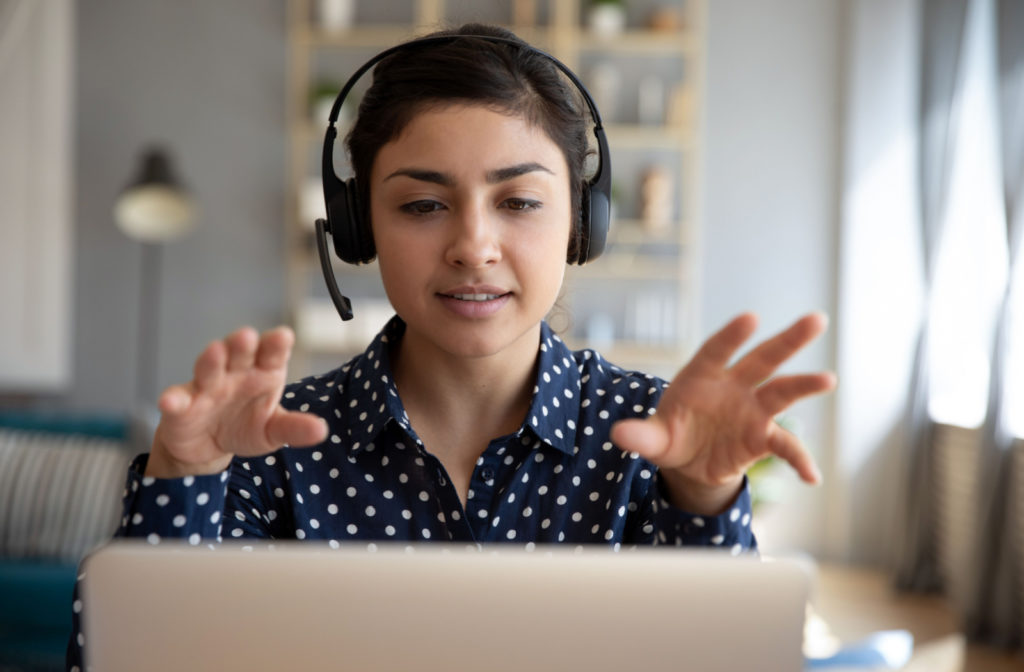 a young woman sits behind a computer wearing a headset and speaking to someone on the computer
